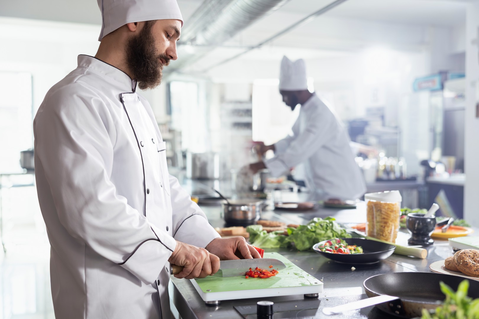 Dedicated restaurant chef chopping a bell pepper in a stainless steel kitchen, preparing tasty ingredients for a gourmet meal. Male cook in a workspace filled with fresh vegetables and utensils.